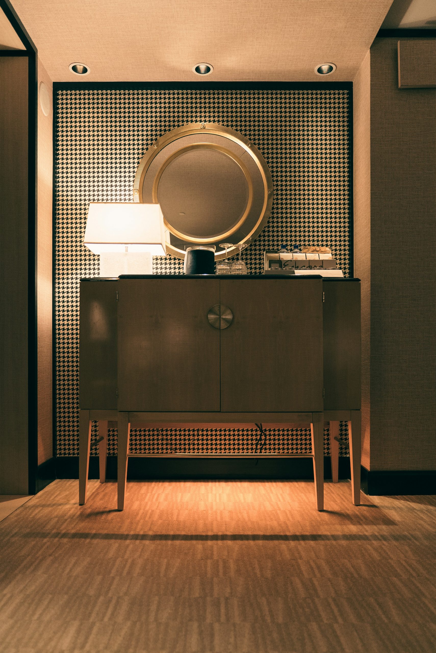 Contemporary Art Deco interior design featuring a large circular brass porthole mirror above a dark lacquered sideboard with houndstooth wallpaper, inspired by SS Normandie ocean liner interiors.